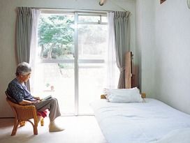 ベッドのそばに置かれた椅子に座って高齢女性が日向ぼっこをしている様子。