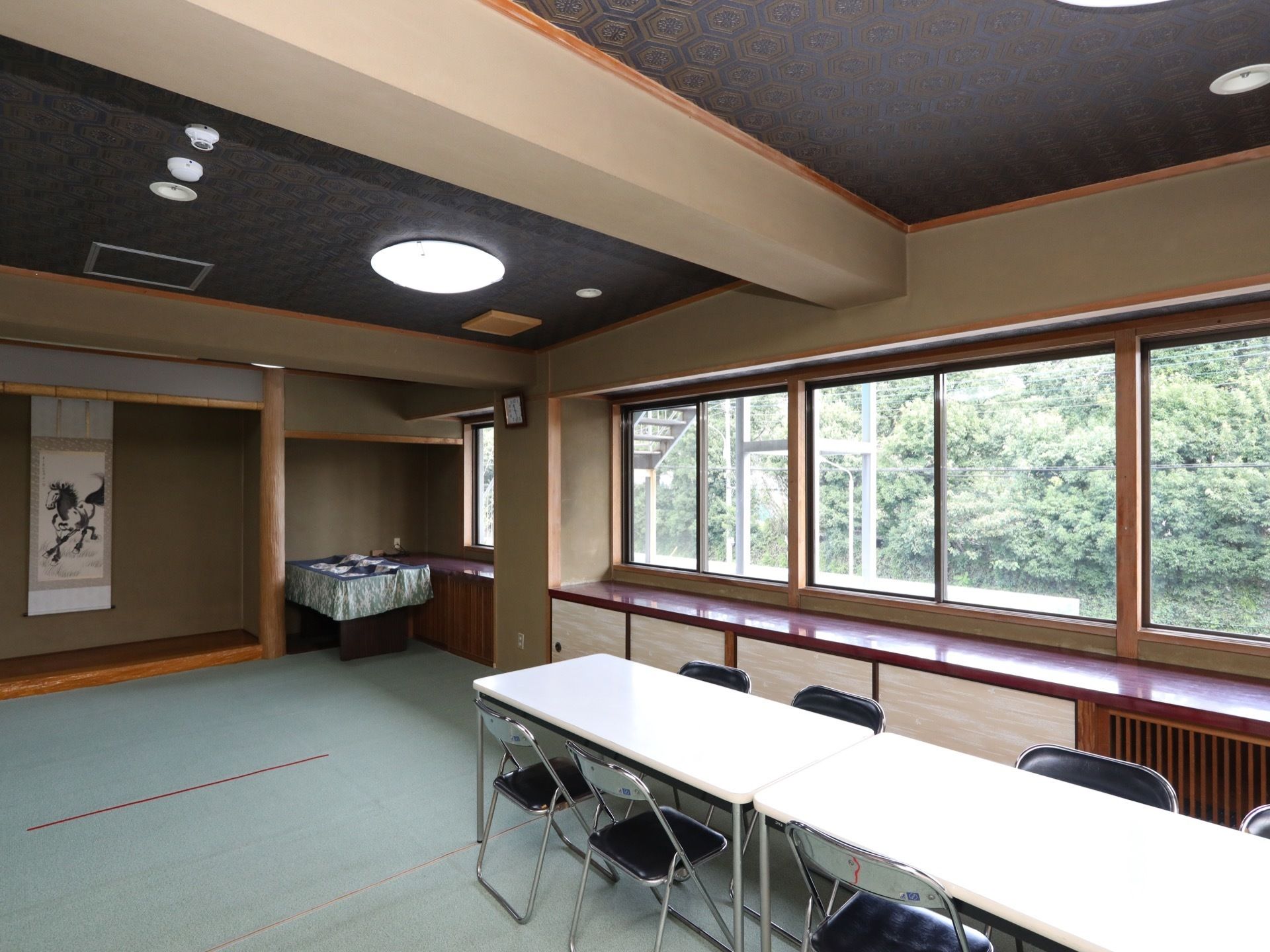 亀甲模様の天井が施された趣のある部屋にテーブルや椅子が並べられている。右側に窓が連続しており、木々の緑が見える。