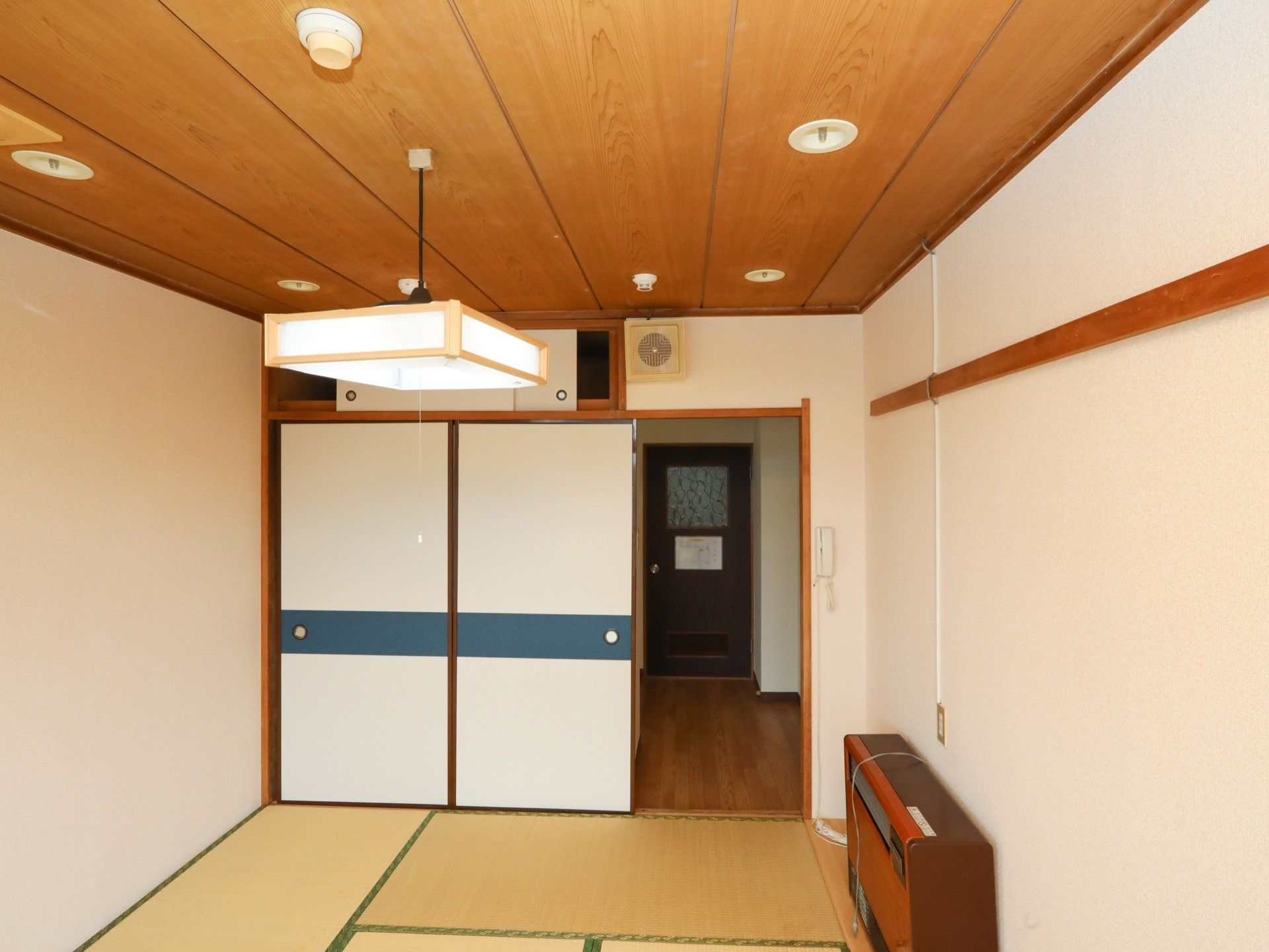 畳が敷き詰められ、四角い照明器具が提げられている和室である。右側の壁際に茶色い暖房器具がある。