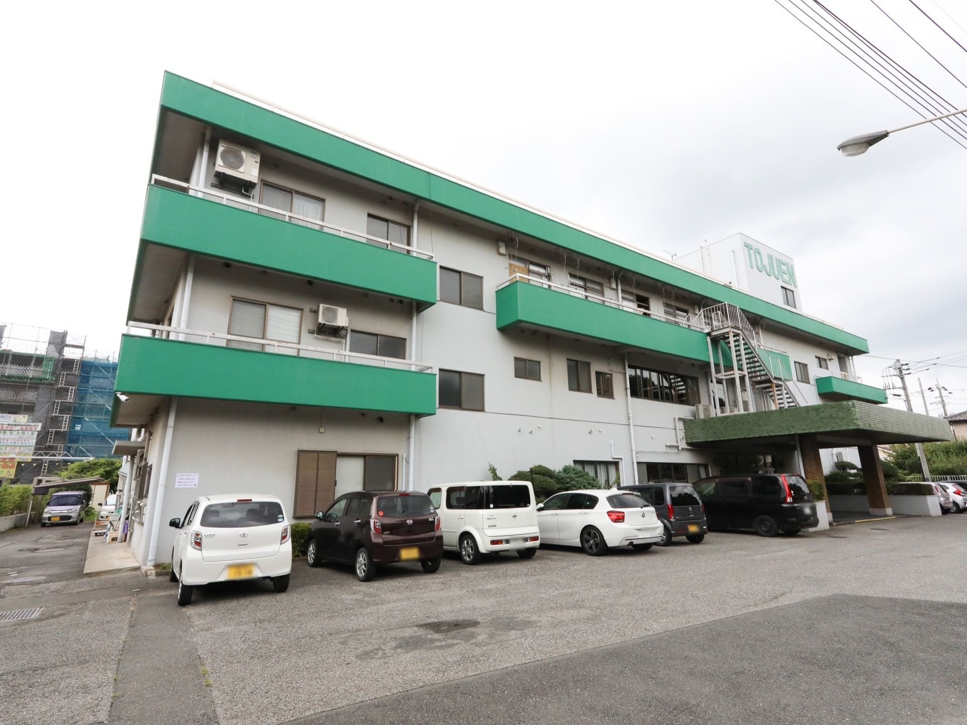 3階建ての建物の周囲は舗装されており、複数の自動車が駐車されている。玄関の前に張り出した屋根の上に非常階段が見える。