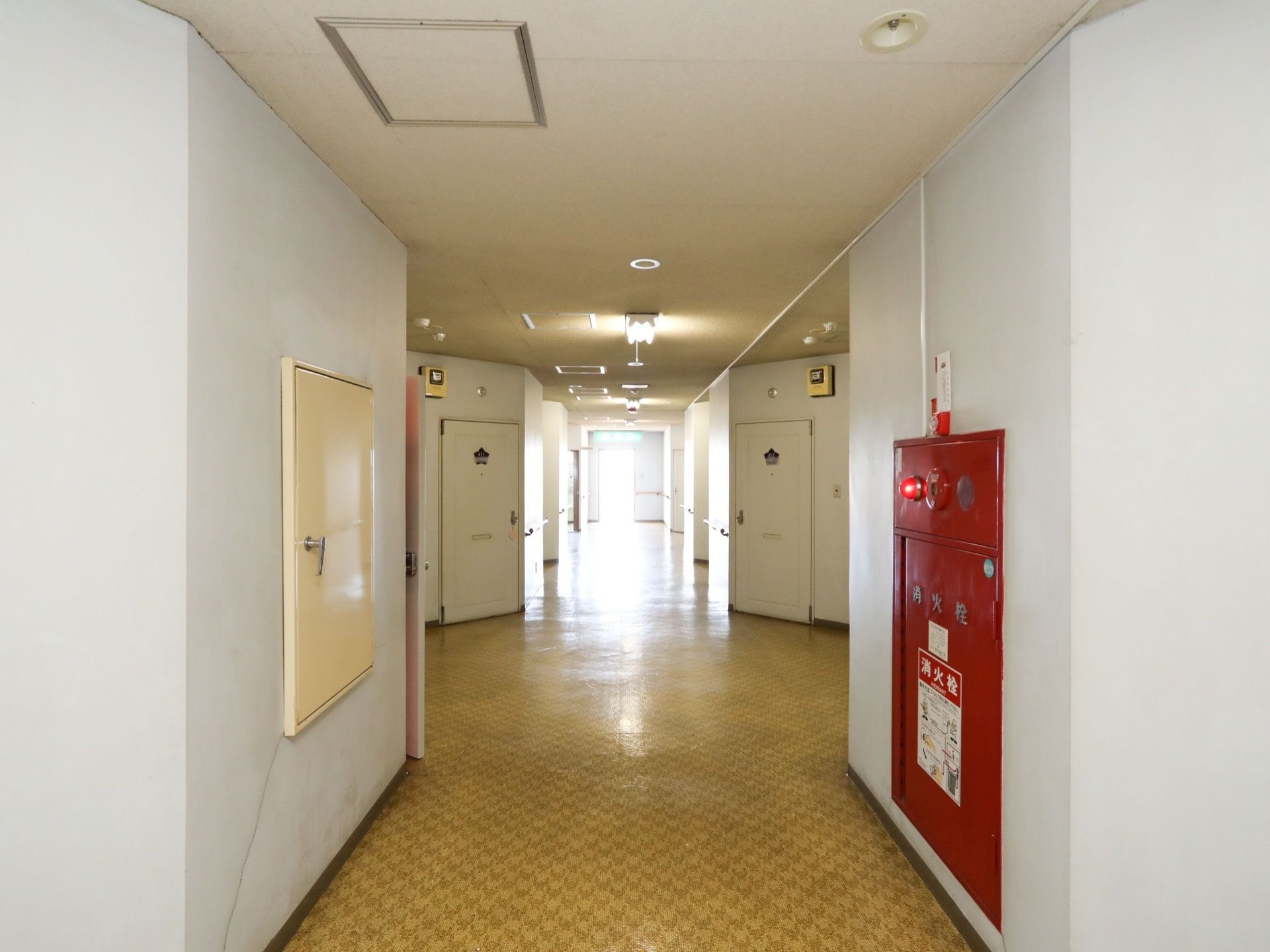 廊下の照明は、長い直管タイプの蛍光灯になっている。壁は無地の白で、床はオレンジで柄のついたラバータイルが使われている。