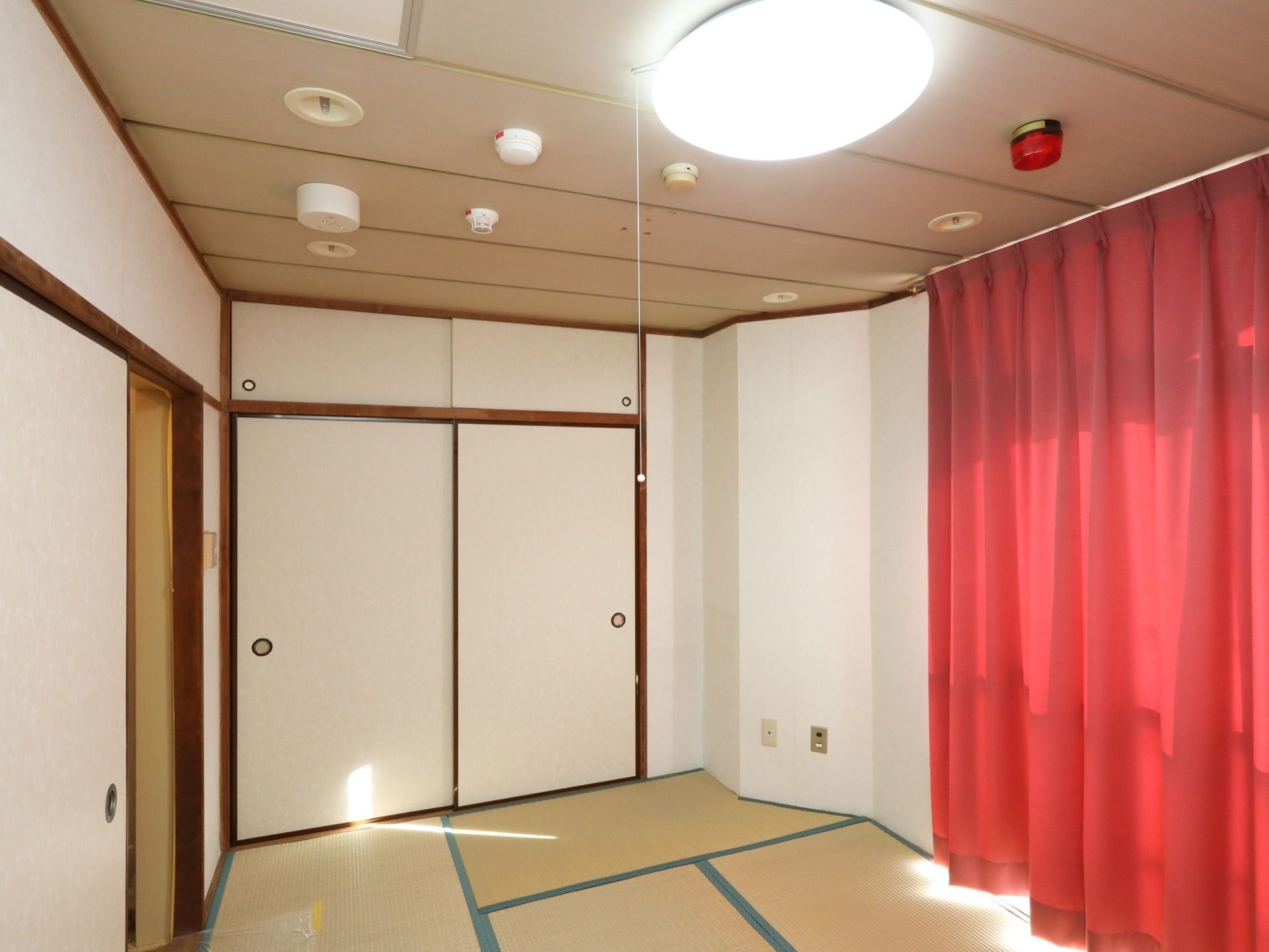 木の板張りの居室の天井には大きく丸型の照明のほか、いくつかのサブランプや火災報知機などが追加で設置されている。