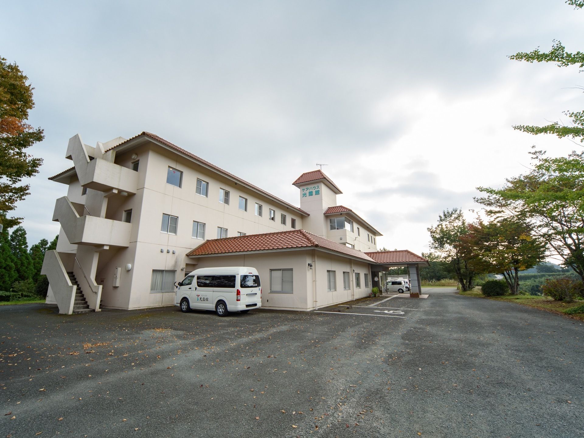 ホワイト調の3階建ての施設外観である。敷地内には車が停められており、車椅子の駐車スペースも完備されている。