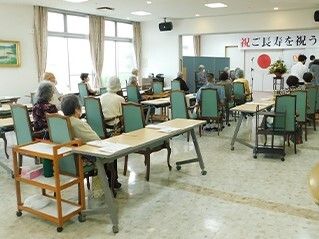 食堂に集まりイベントを楽しんでいる高齢者たち。敬老の人の垂れ幕がかかり、前を向いて座っている。