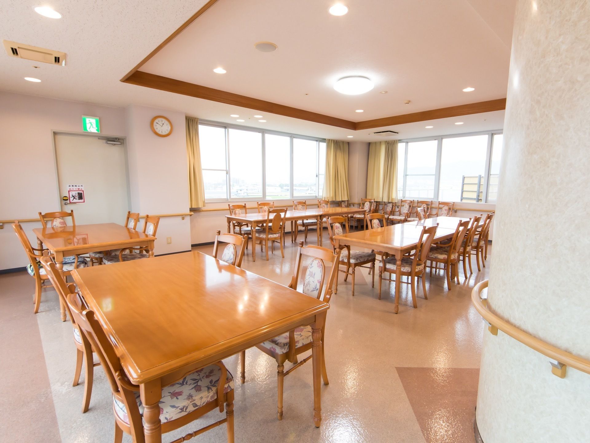 窓に囲まれて明るい食堂がある。テーブルセットがたくさん並んでいて、壁には手すりがある。