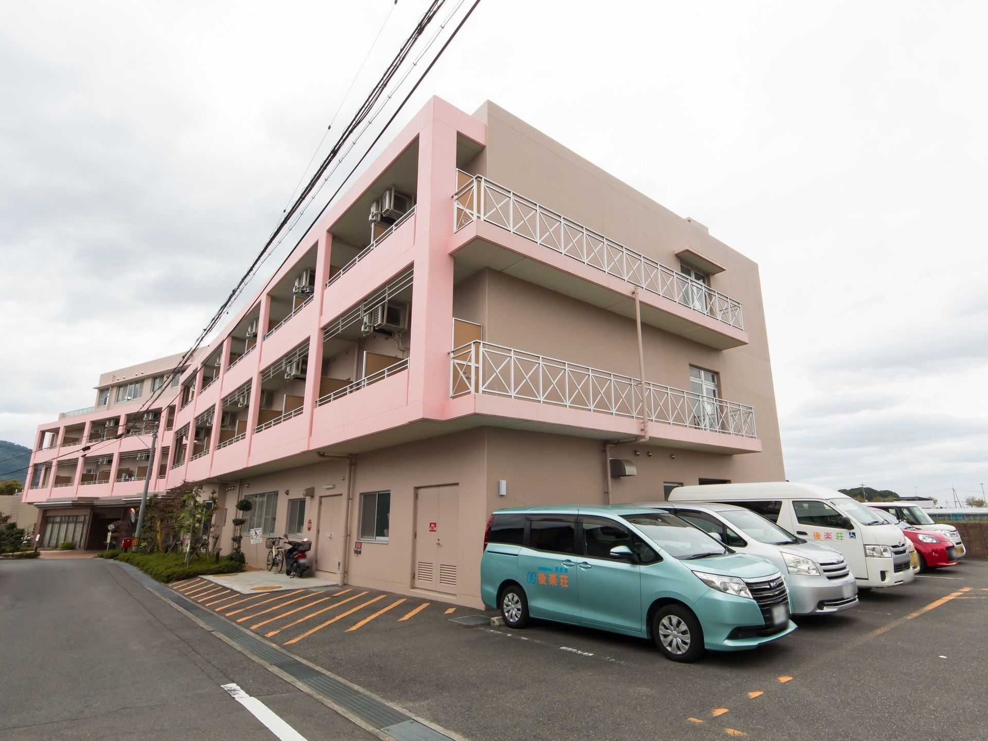 ４階建てのマンションが建っている。外壁は淡いピンク色で、建物の横には駐車スペースがあり、車が5台ほど停まっている。