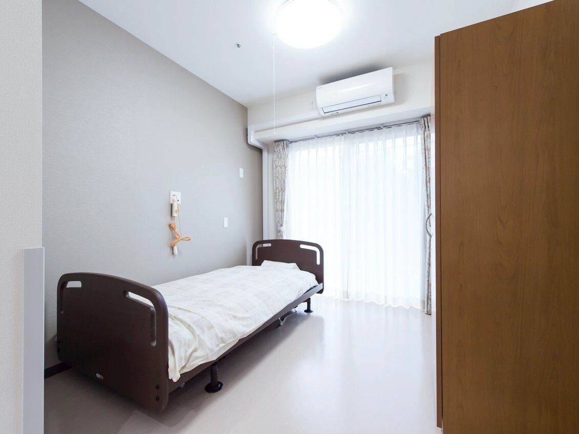 緊急通報装置が付いている部屋には寝具付きのベッドが置かれ、窓にはカーテンが付いている。