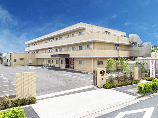 3階建ての建物はベージュ系の外壁で駐車場も付いている。門のそばには草木が植えられている。