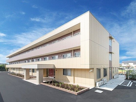 ベージュ系の外壁で敷地内には駐車場があり、3階建てで各フロアにベランダがある。