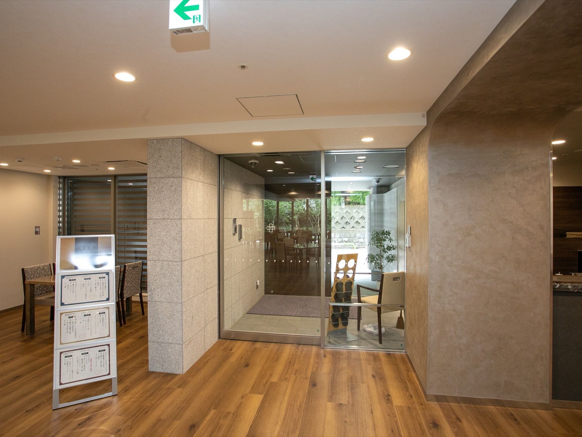 木目のフローリングの床に表示板が置かれている。ガラスの自動扉の向こう側に椅子とスリッパラックがある。