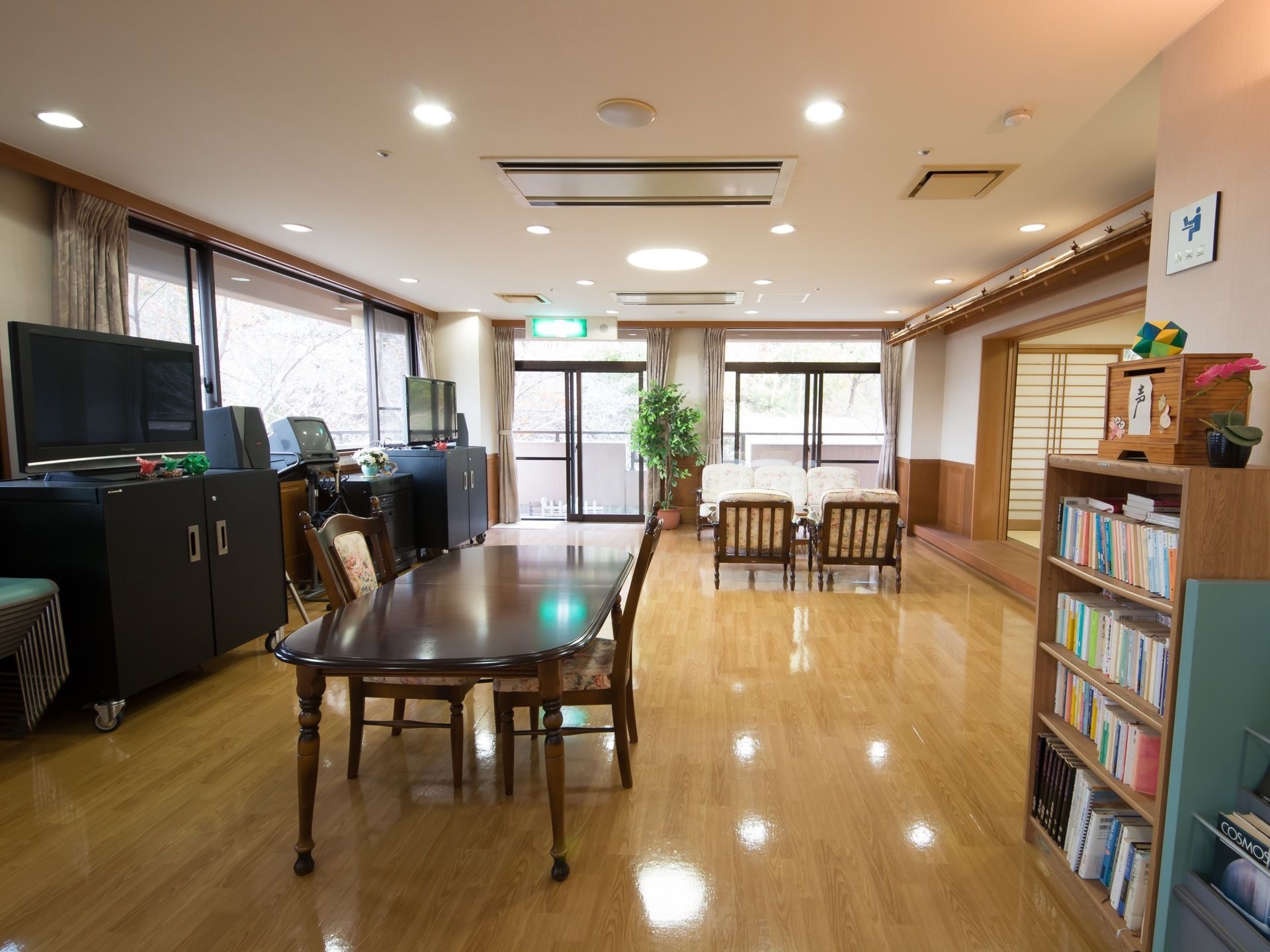 広く開放的な空間にダイニングテーブルとソファーが置かれている。テレビが２台設置されており、本棚には本が並んでいる。