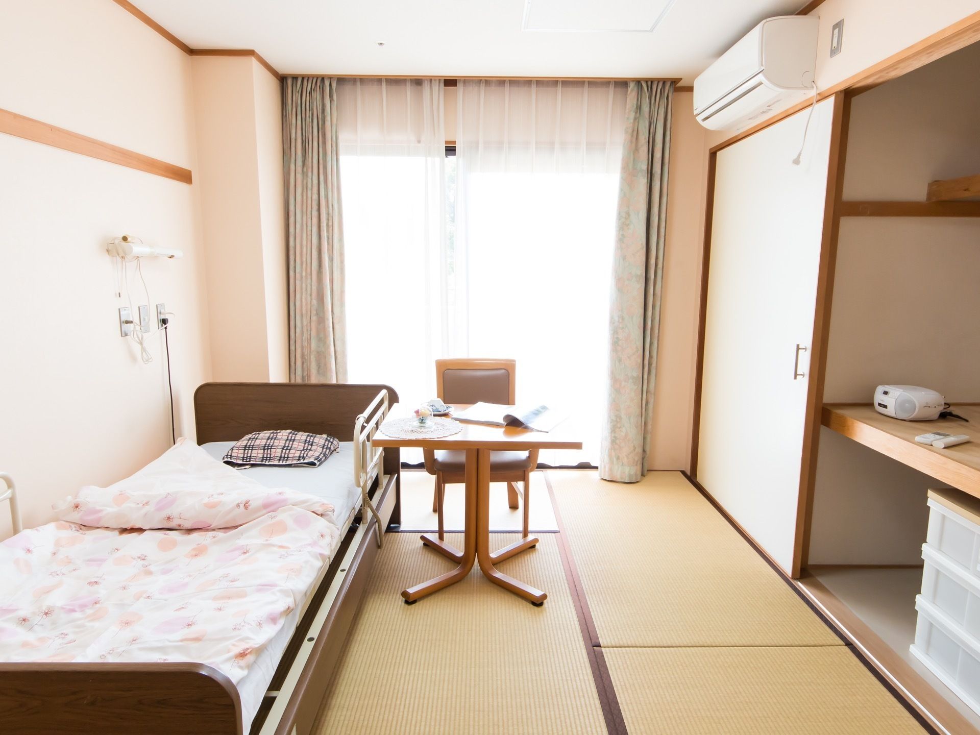 掃き出し窓が設置されており、日差しが降りそそいでいる。畳が敷かれた和室タイプの居室。ベッドやテーブル、椅子が置かれてある。