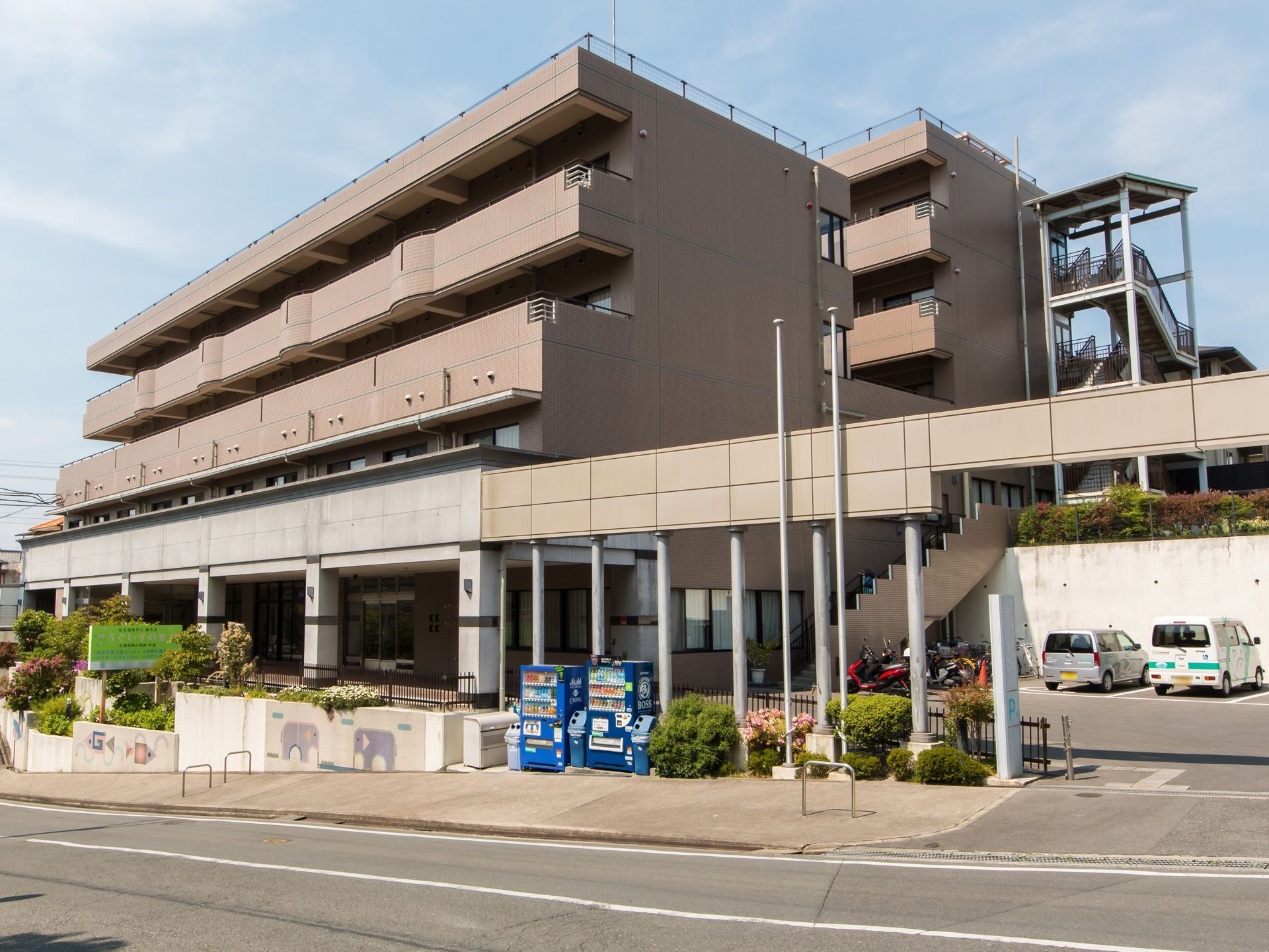 落ち着いた色調にまとめられた外観である。目の前は道路が走っており、歩道が完備されている。青色の自動販売機もある。