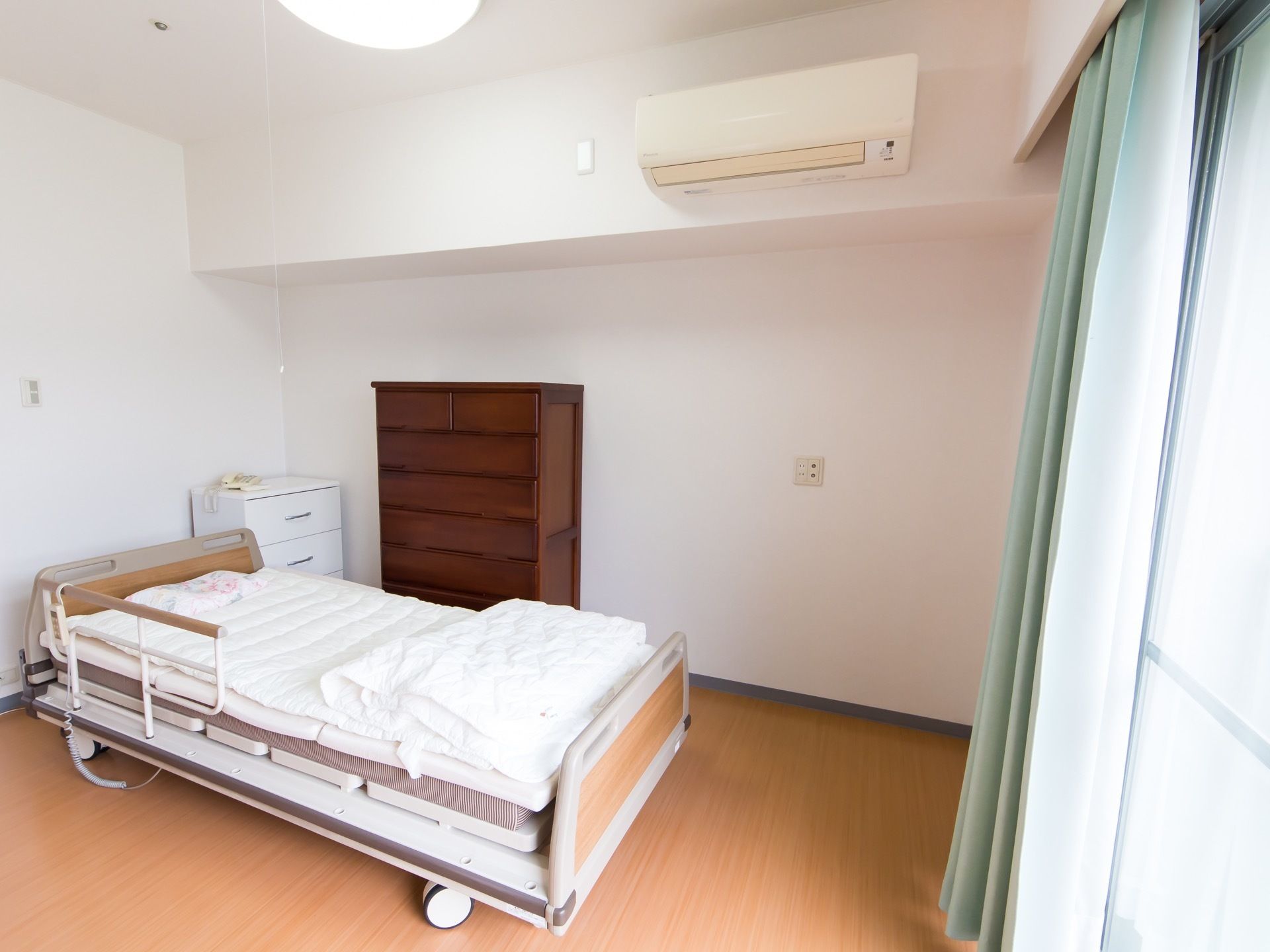 真っ白な壁が清潔感を生み出している居室である。介護用ベッドやタンス、エアコン、カーテンが完備されている。