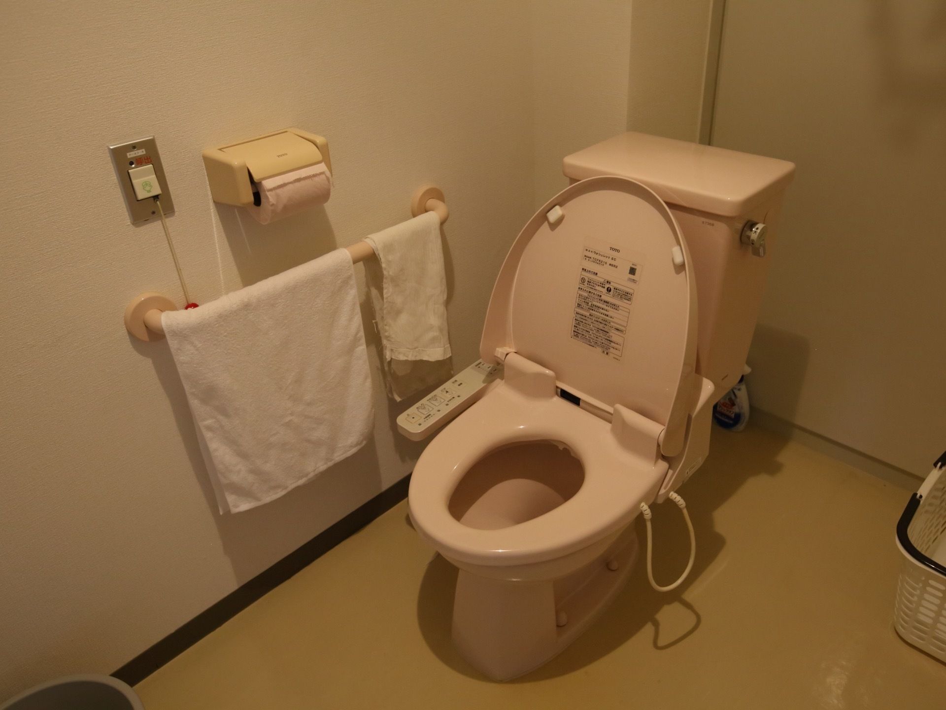 温水洗浄便座器が完備されたトイレである。壁には、手すりと緊急呼び出しブザー、ペーパーホルダーが設置されている。