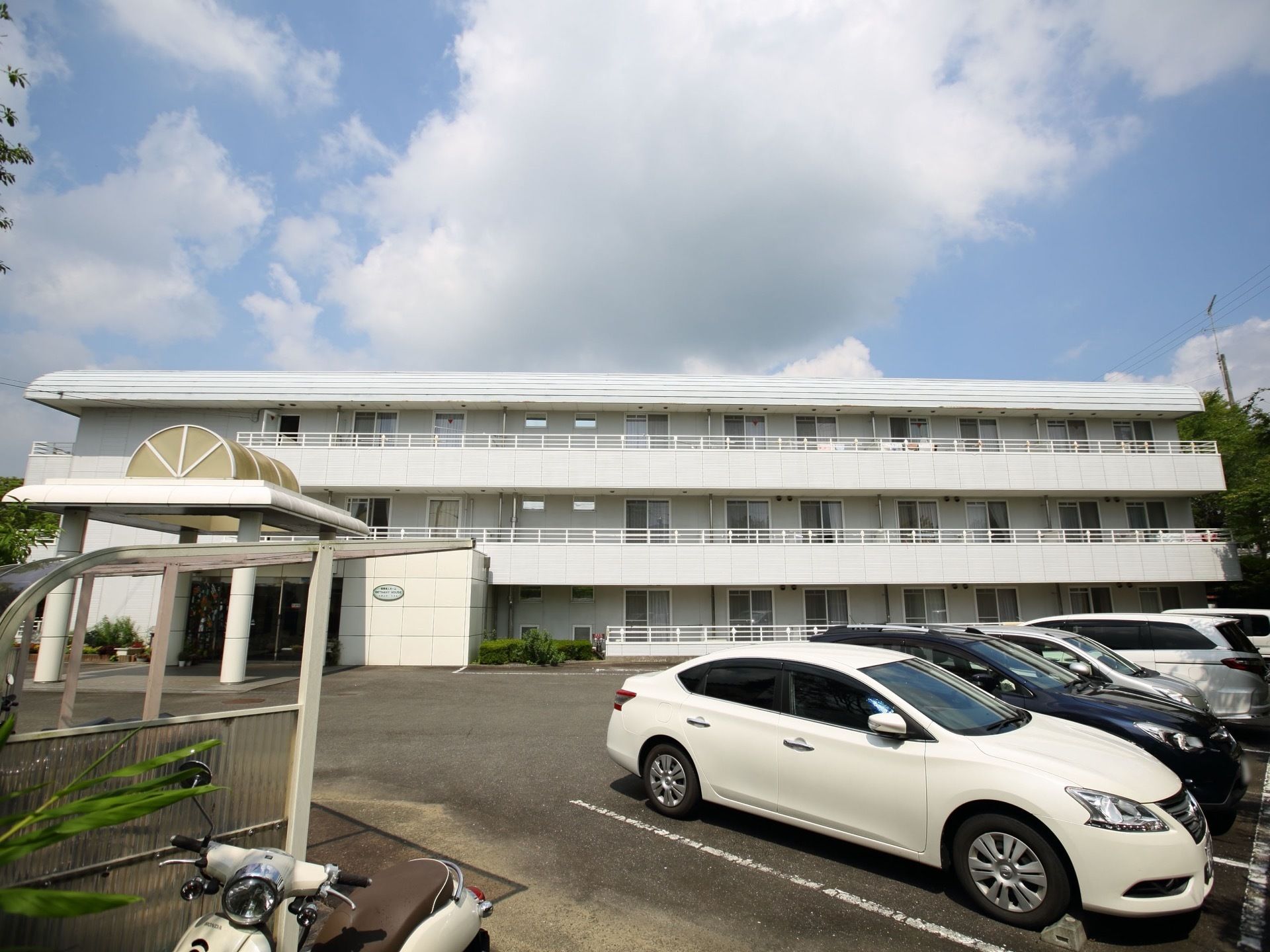 それぞれの居室にベランダが完備された、白い外観の3階建て施設である。駐車スペースが設けられ、乗用車が数台停車している。