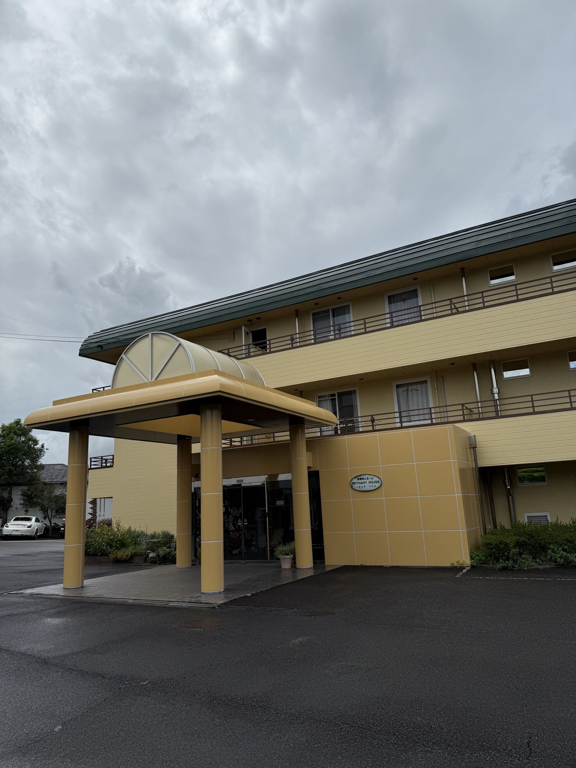 それぞれの居室にベランダが完備された、白い外観の3階建て施設である。駐車スペースが設けられ、乗用車が数台停車している。