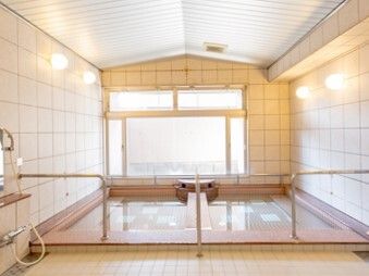大きな窓が付いた大浴場には大きな湯船が設置され、手すりが浴槽を囲むようについている。左側にはシャワーも付いている。