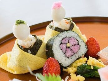 赤い盆の上にのせられたひな寿司。ひな人形の形をした寿司の周りにはフルーツやおかずが盛られていて、色も鮮やか。