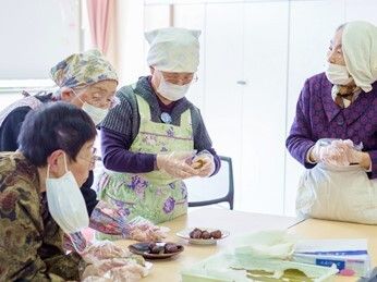 エプロンや三角巾、マスクをつけた高齢女性たちが和菓子を作っている様子。真剣な表情で何かをこねている。