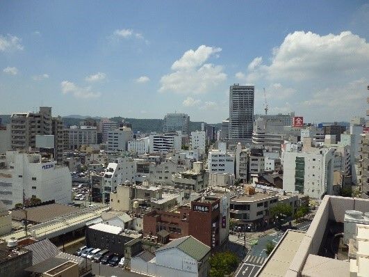 たくさんのビルが建ち並んでいる街並み。道路には車が通り、人が行き交っている姿が見える。遠く向こうには山々もある。