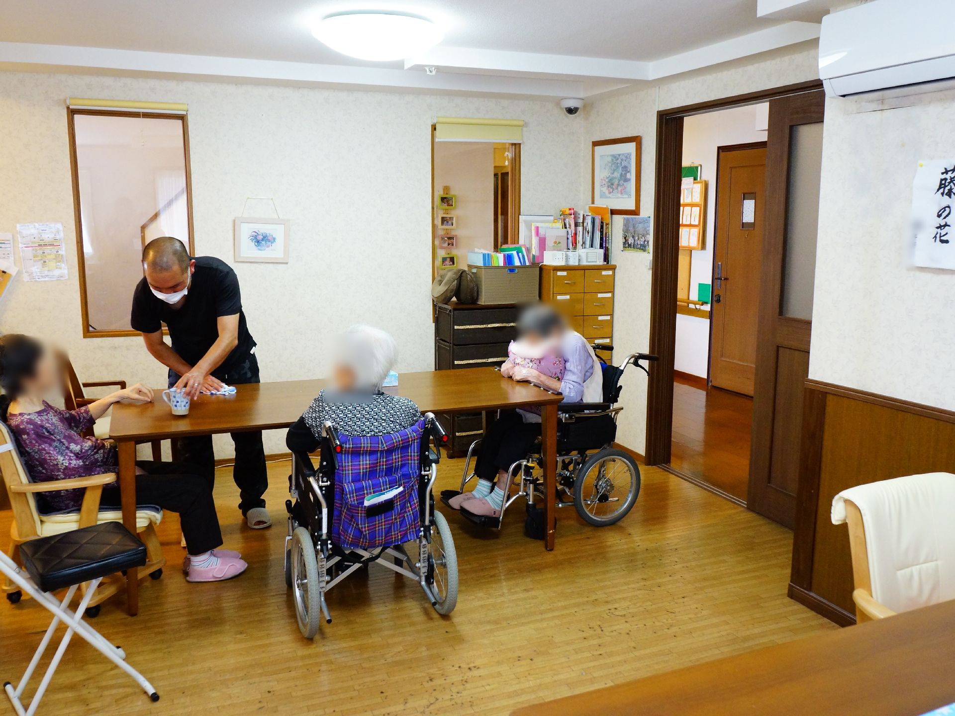 ダイニングテーブルには女性の入居者様が3人座っている。二人は車いす、ひとりは椅子に座っている。職員が一人ついている。