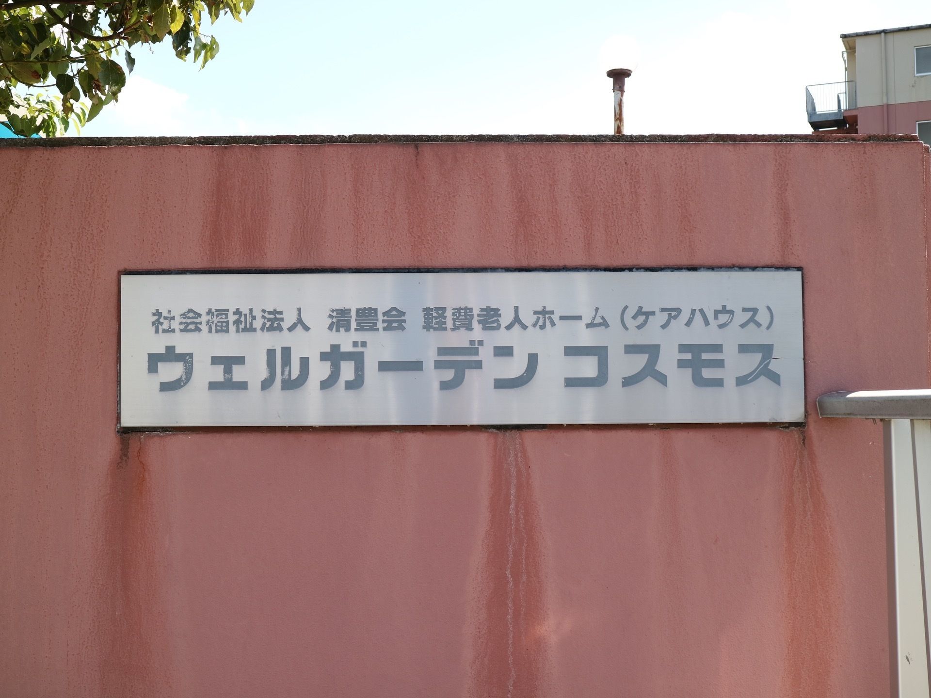 薄赤色の壁に横長の銘板がある。そこに、運営母体と提供サービス、その下に大きな文字で施設名称が表示されている。
