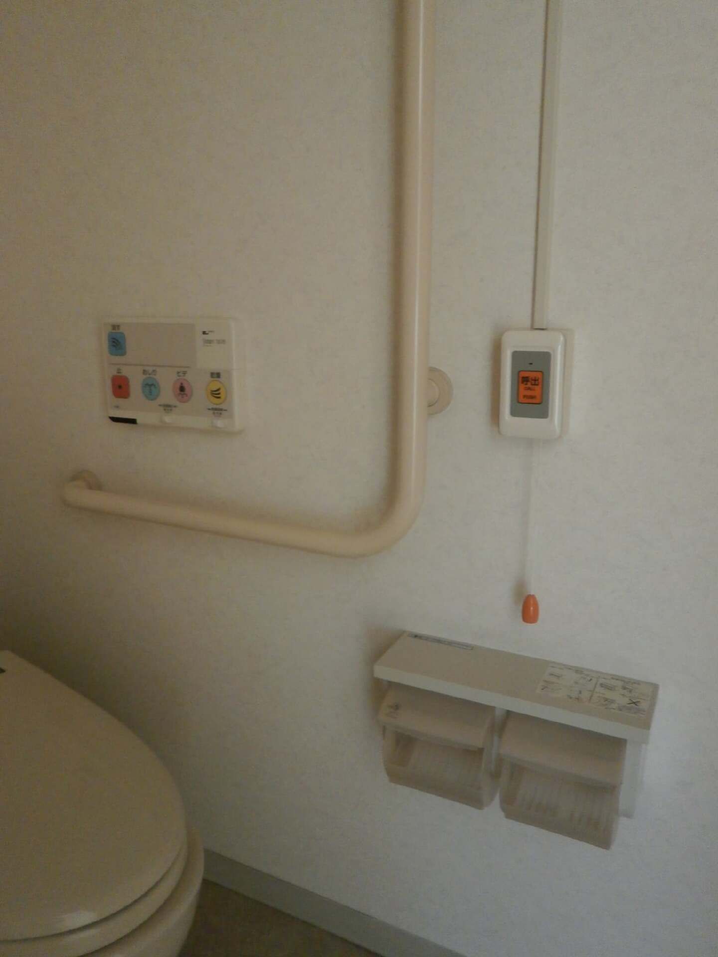 操作パネルやエル字型の手すりやオレンジの呼び出しボタンが付いたトイレ。トイレットペーパーホルダーは二連式。