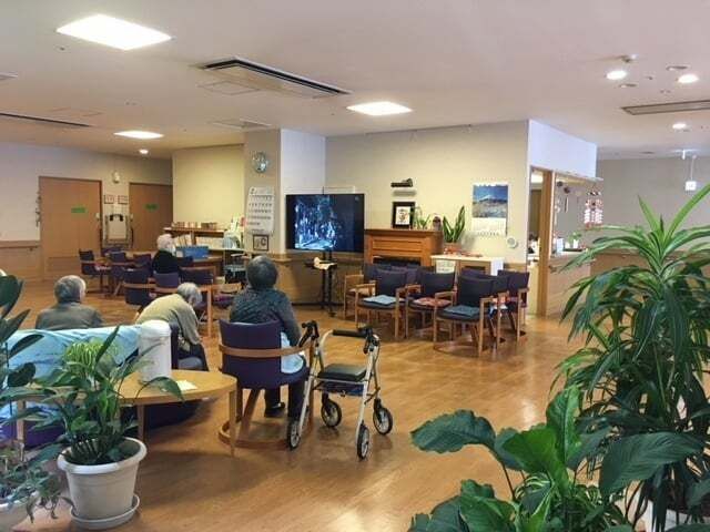 複数の高齢者が集まっている共有スペース。観葉植物が飾られ、テレビも置かれている。