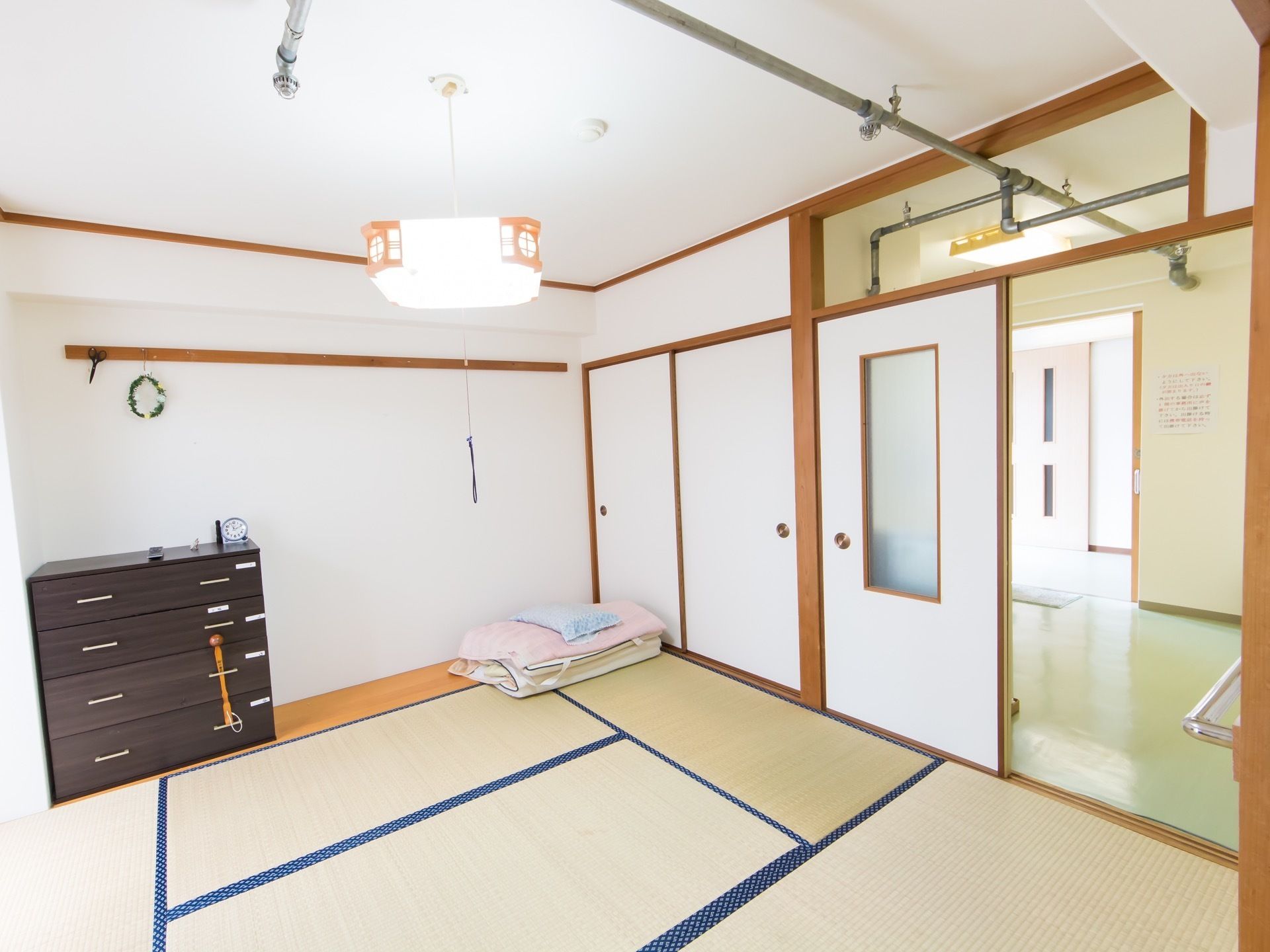和室に引き出し型の収納家具や折りたたまれた布団が置いてある。部屋の入り口の引き戸には、大きな四角い窓ガラスが付いている。