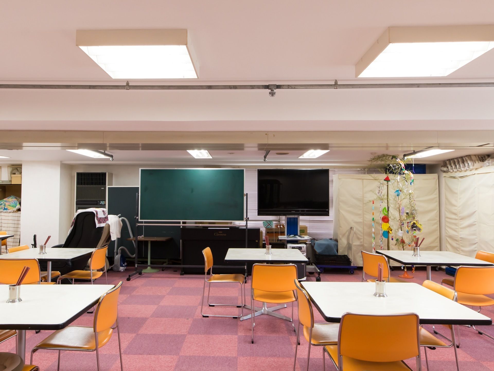 市松模様のカーペットが敷き詰められた広間に、複数のテーブルやパイプ椅子が置かれている。正面に液晶テレビと黒板がある。