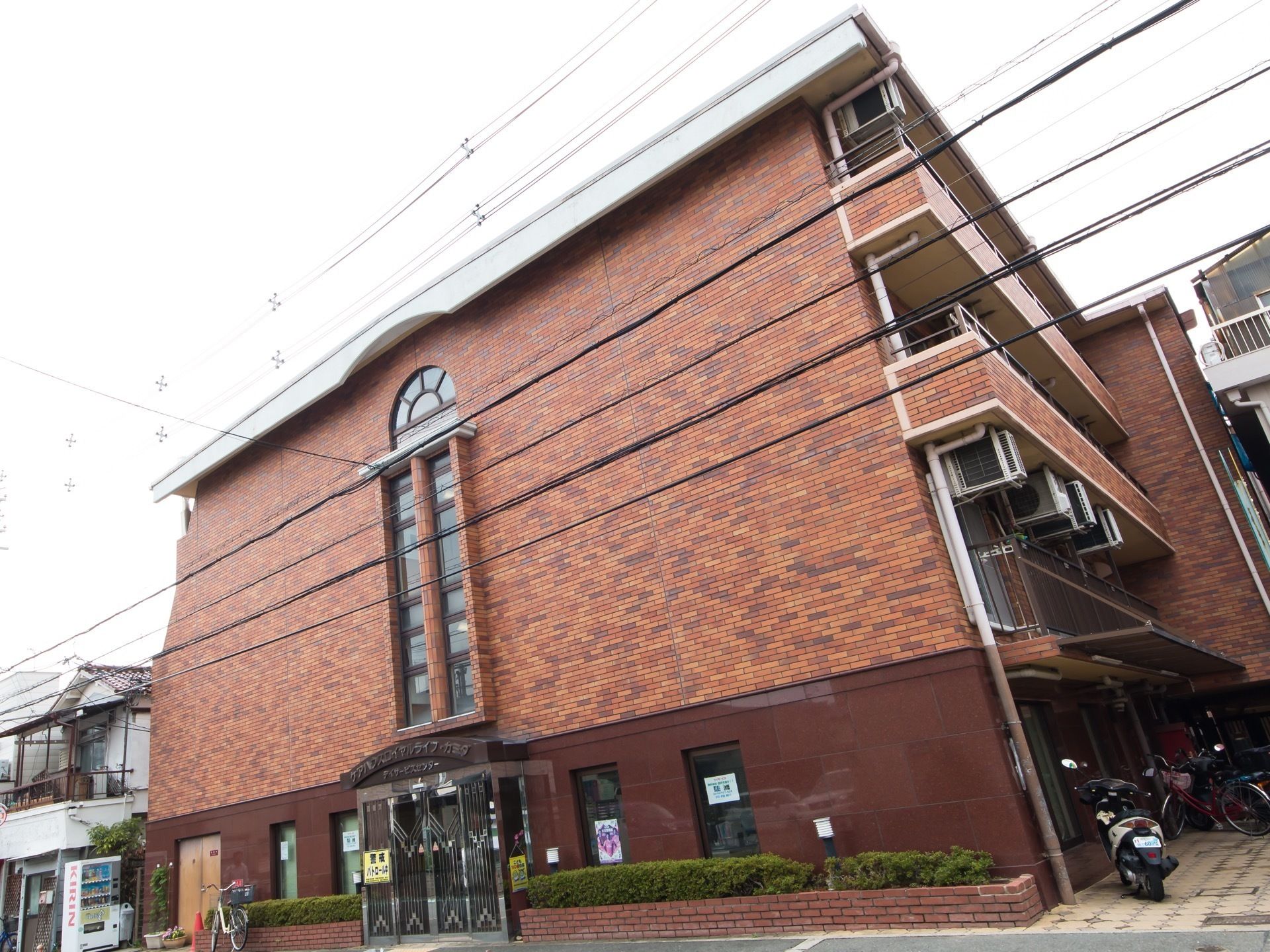 レンガ調の地上4階建ての施設が建っている。右側がタイル張りの駐輪スペースになっていて、自転車やオートバイが停められている。