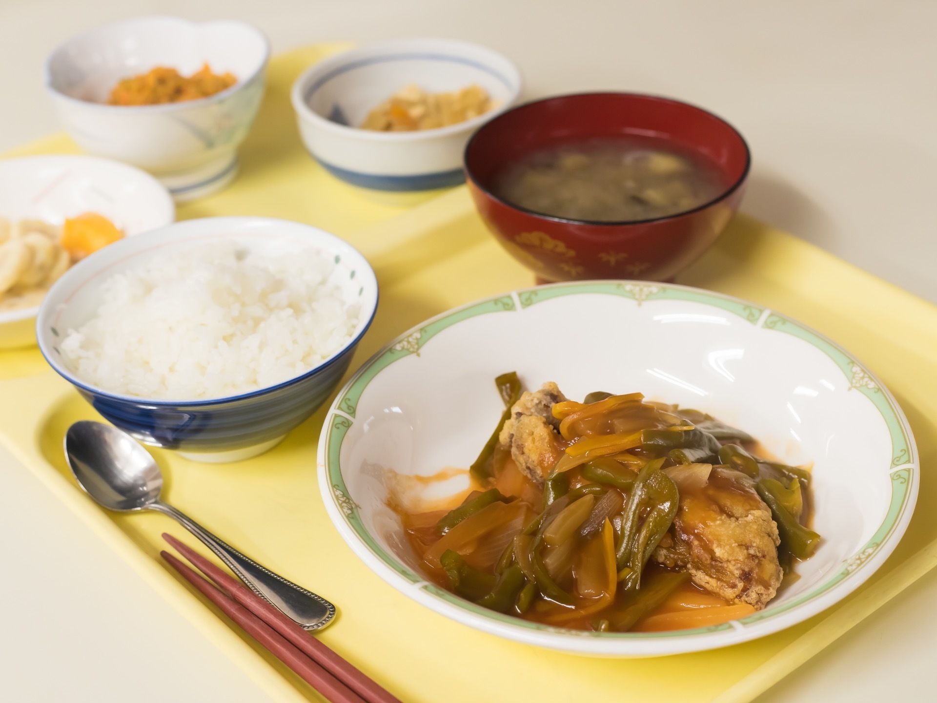 ご飯と味噌汁のほかに、あんのかかった揚げ物と副菜2種、果物の小鉢がトレイに載せてある。箸とスプーンが用意されている。