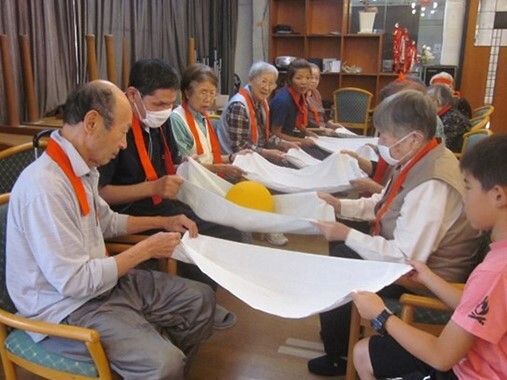 対面式に椅子に座って白い布をペアで持ち、隣のペアにボールを渡す競技を行っている高齢者たち。