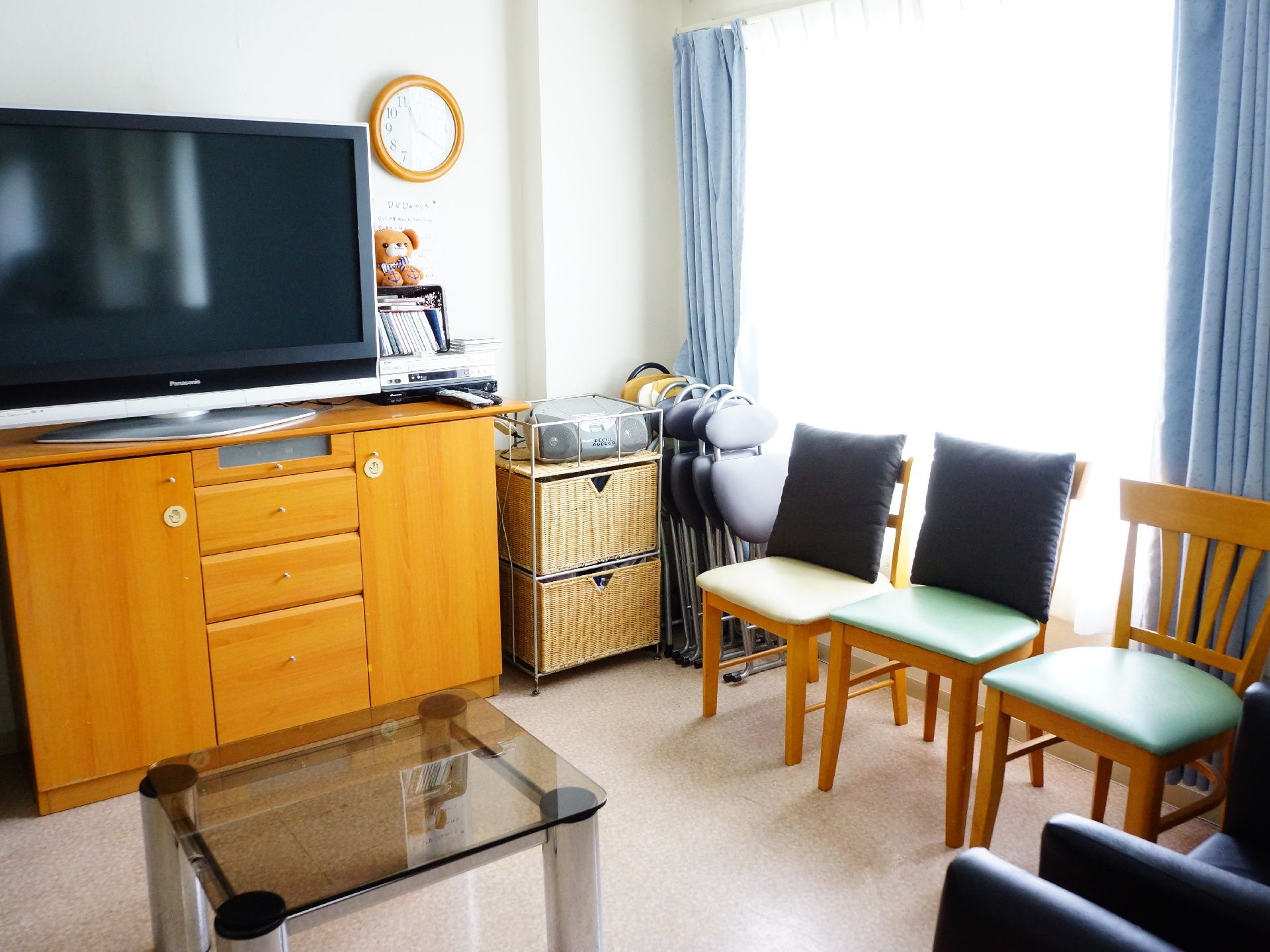 木目調の戸棚の上に大型のテレビが設置されている。木目調の椅子が窓際に並び、手前にはガラス製のテーブルが置かれている。