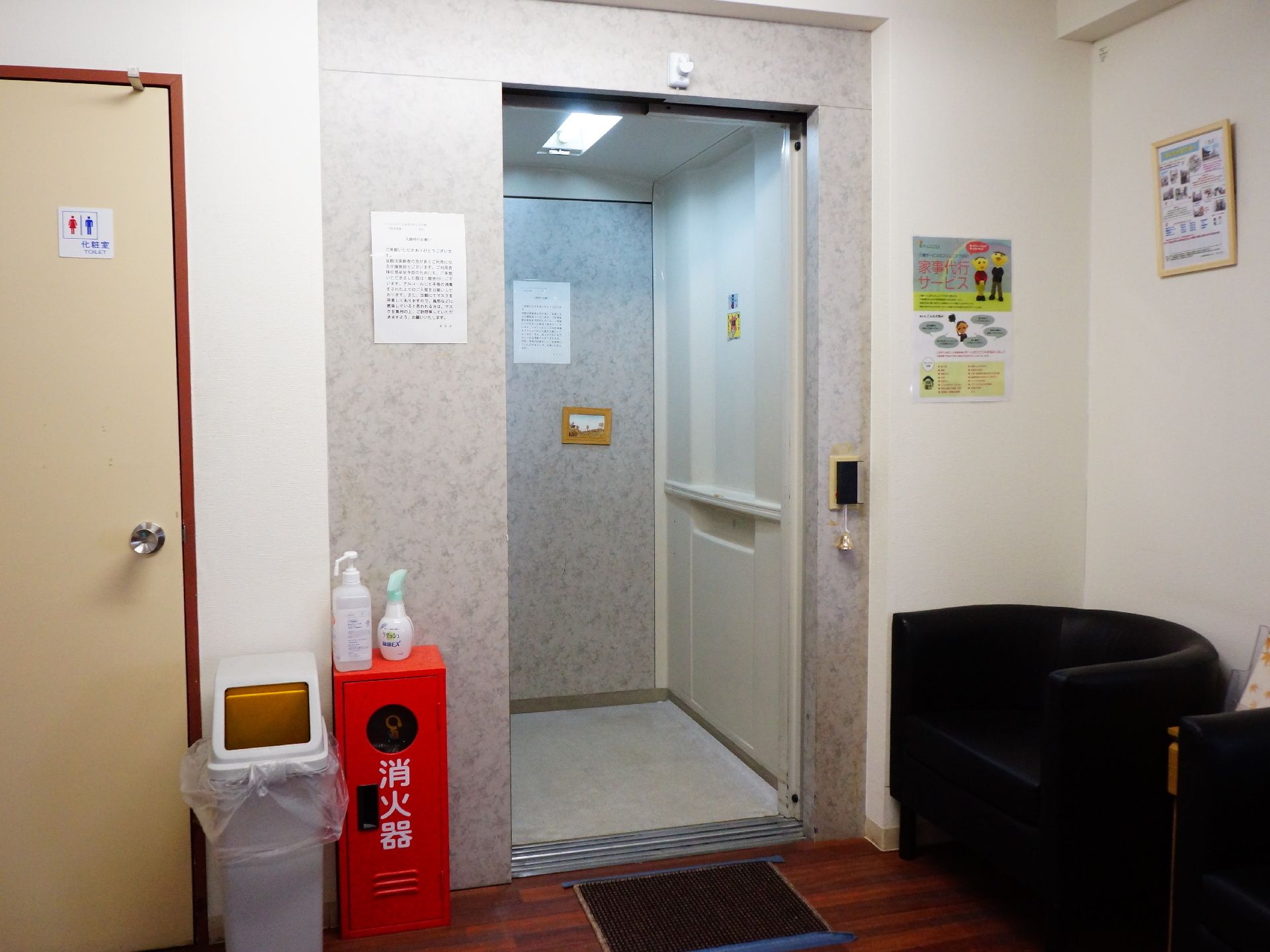 エレベーター周りはグレーで統一されており中には張り紙がみえる。右側に2つの半円形の黒いソファが置かれ、左側に消化器などが設置されている。
