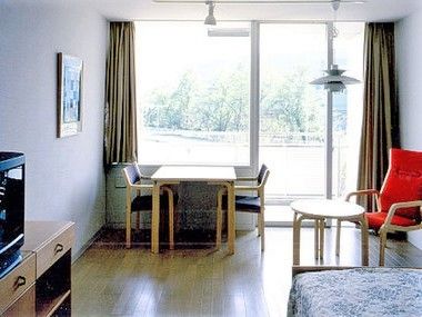 広めのワンルームタイプの部屋にテーブルと椅子、テレビ、ベッドが置いてある様子