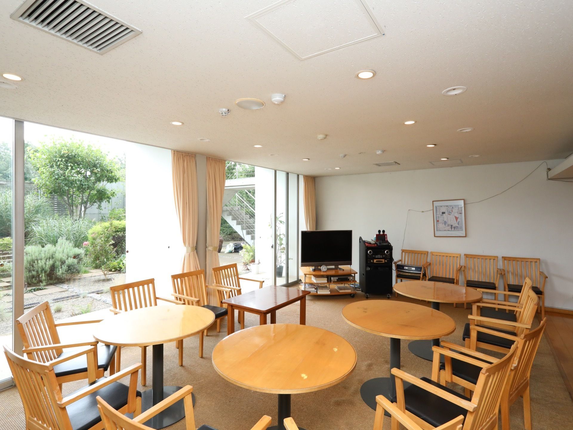大きな窓が設けらた談話室である。複数の椅子とテーブルが置かれている。壁際にカラオケ機器とテレビがある。