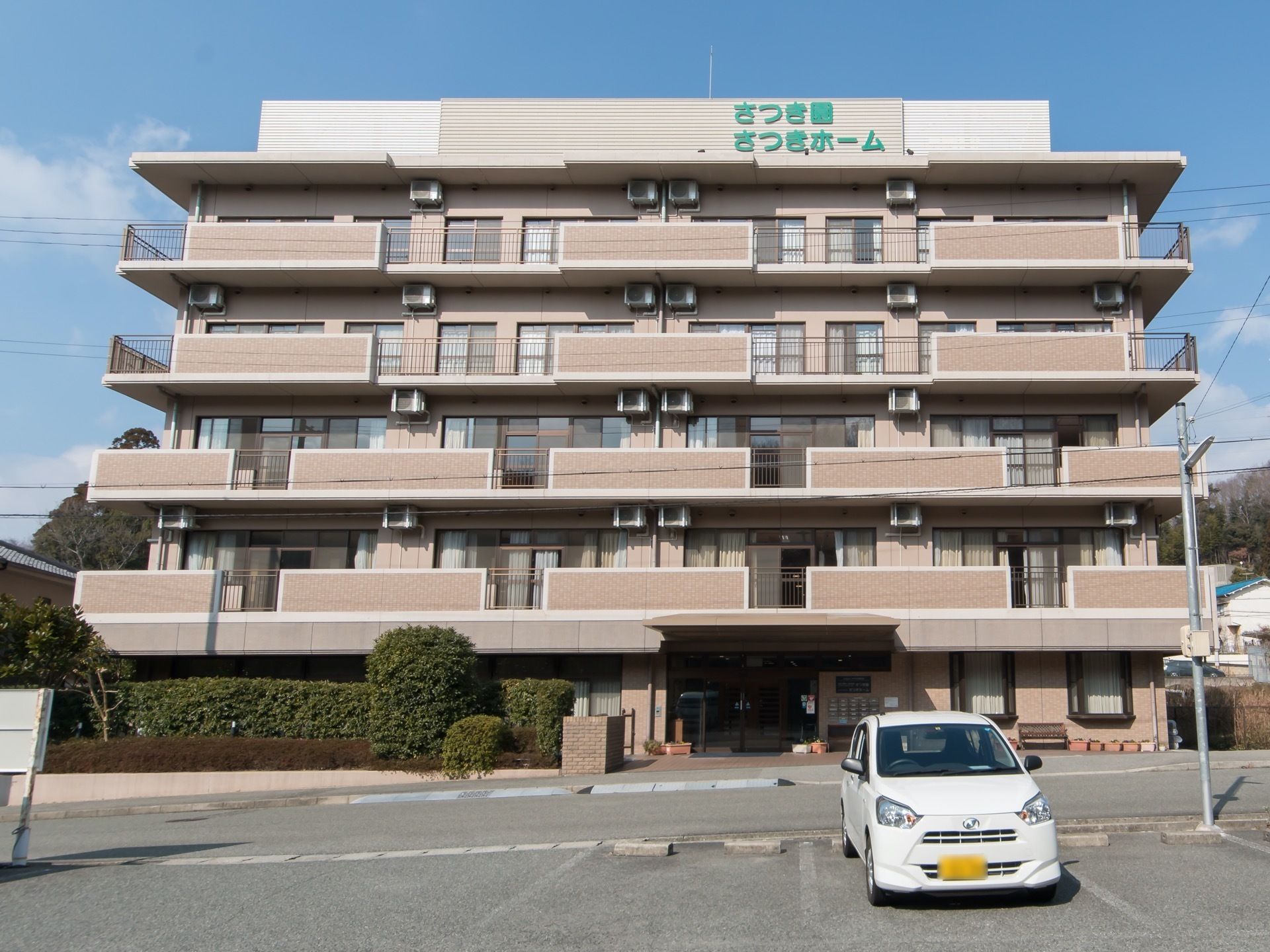 青空の下に５階建ての建物が立っているようすである。最上部には、施設名が書かれており、建物の前には車が停まっている。