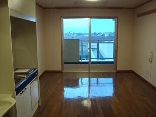 フローリングの部屋にはIHクッキングヒーター付きのキッチンや緊急通報装置、大きな窓があり、窓にはカーテンはない。