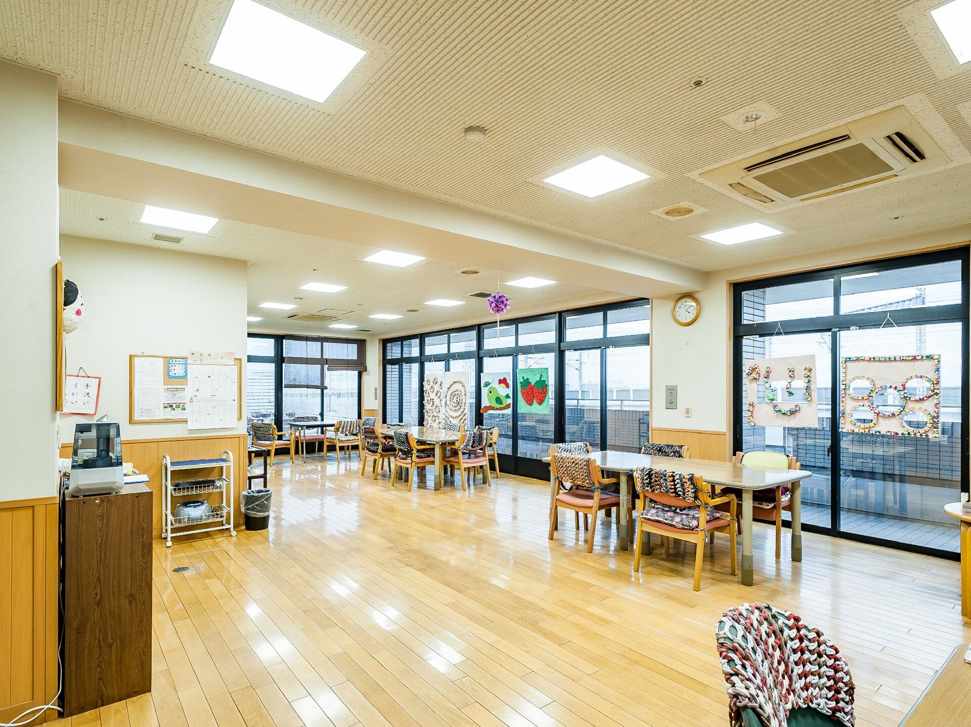 白衣を着た女性スタッフと高齢女性たちがソファを囲んで楽しそうに話している。スタッフは両隣の女性たちと手をつないでいる。