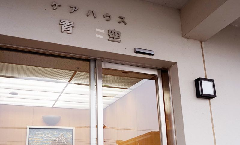 ケアハウスの名前が書かれた玄関の外壁。ガラスのドアが設置されていて、照明も付いている。
