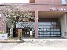 ハウス名が外壁に書かれたブラウン系の建物の前には木や花が植えられたスペースがある。