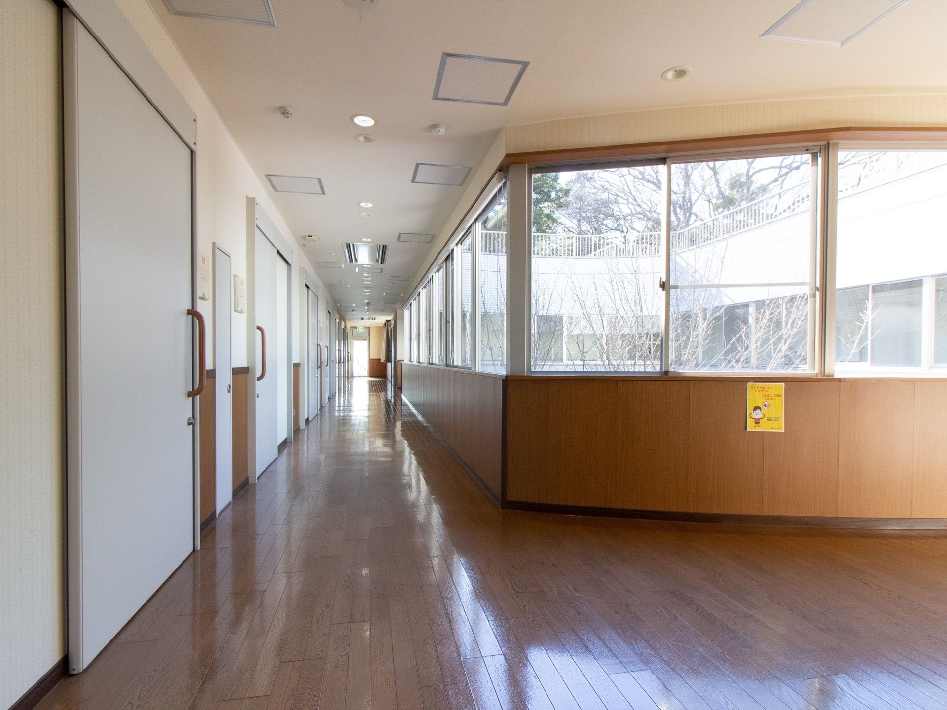 居室の白いスライド式のドアが並んでいる、茶色のフローリングの廊下。片側が一面窓になっており、中庭が見える。