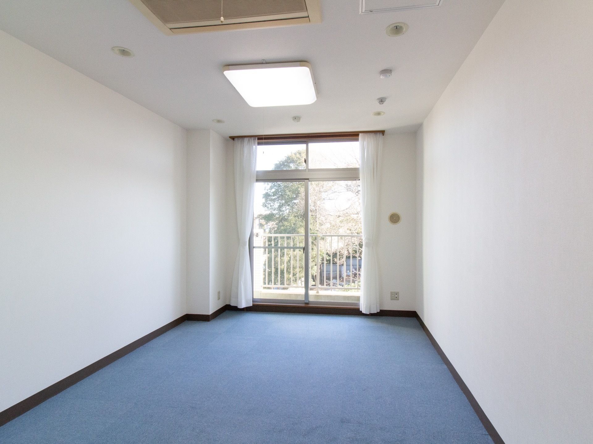 白い壁に青い絨毯が敷かれた居室がある。大きな窓があり、カーテンが掛けられている。柵のついたベランダが設置されている。