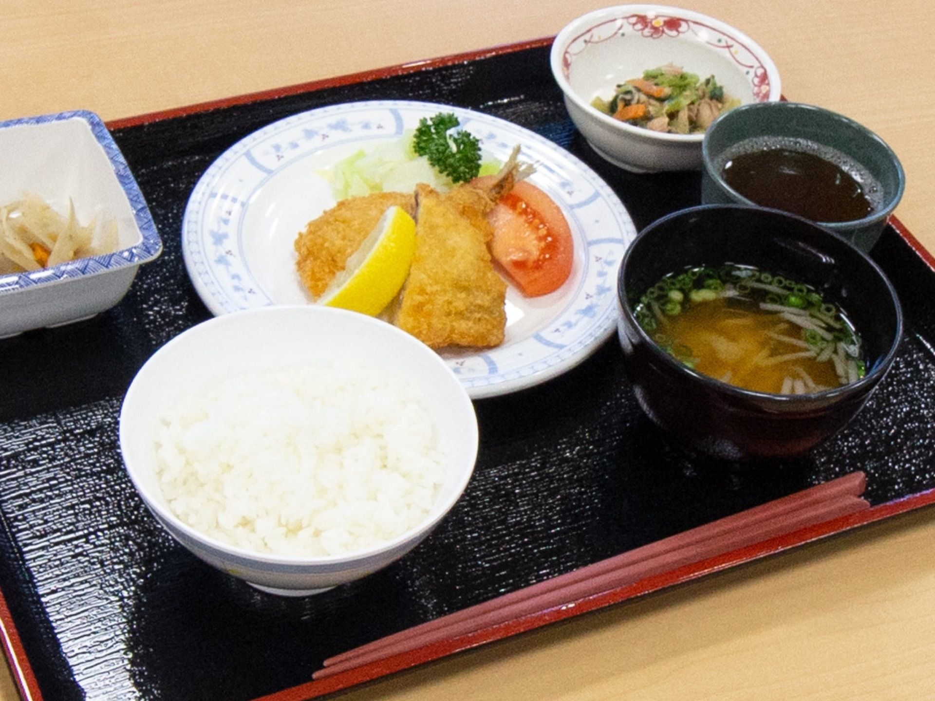 黒いお盆に食事が乗っている。魚のフライ、きんぴら、あえ物、ご飯、味噌汁、お茶がそれぞれ個別の皿に盛られている。