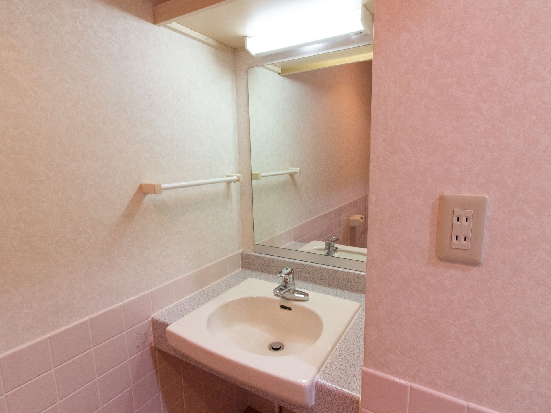 居室の洗面スペースである。ピンク色のタイル張りの部屋にトイレも設置されている。大きな鏡と照明がついている。
