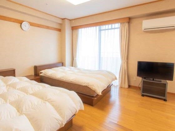 寝具がセットされたベッドが二台置かれたフローリングの部屋には、テレビやカーテン付きの窓、時計などがある。