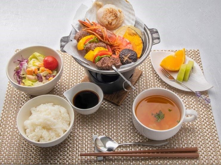 エビや貝などの魚介類が入った鍋料理に白いご飯、スープ、野菜の和え物などが並んでいる。カットフルーツもある。