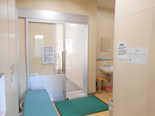 共同浴室の脱衣所に、薄緑色の長いベンチが置かれている。洗面台は自動水栓で、シンクの下に木の丸い椅子がある。